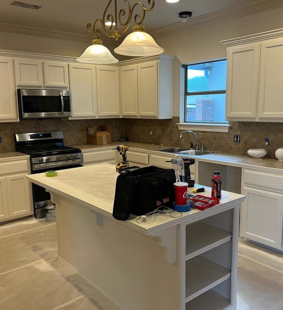 A spacious kitchen renovation by Francise Construction Services in Baton Rouge, with white cabinetry and island, topped with light countertops, illuminated by a classic chandelier, amidst ongoing construction work.