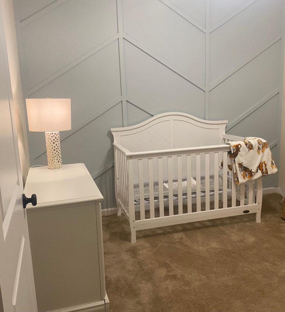 A cozy and serene nursery room designed by Francise Construction Services in Baton Rouge, featuring a white crib against a light gray paneled wall, with a white nightstand and a lamp, complemented by a soft brown carpet.