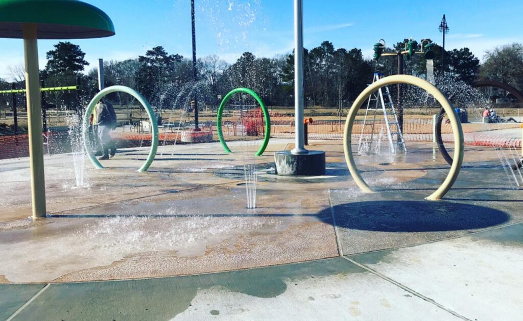 Vibrant water park playground with multiple water jets and arches, including green and yellow hoops, on a concrete surface with children playing, under a sunny sky with trees in the distance.