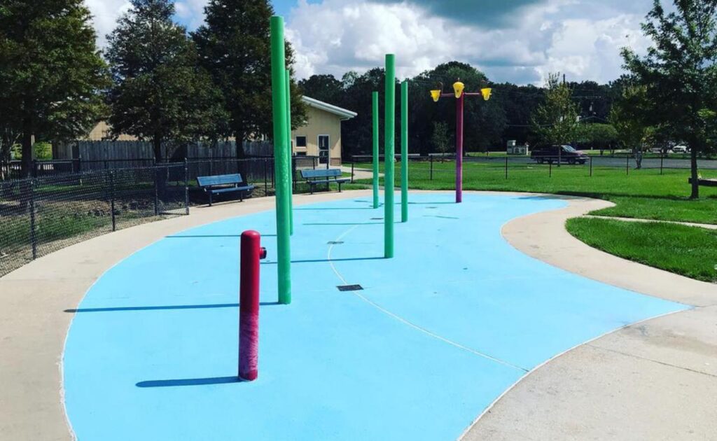 Colorful outdoor splash pad park with blue anti-slip surface and multiple vibrant water spray poles, surrounded by greenery and seating benches under a sunny sky.